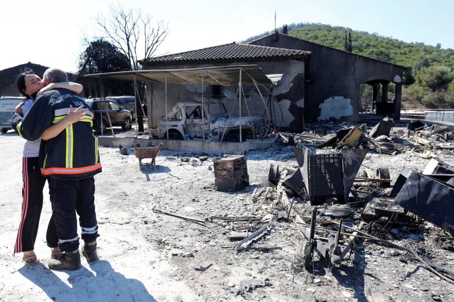 Uma mulher abraça um bombeiro perto dos restos carbonizados de veículos que foram destruídos pelo fogo, em um estacionamento de Bormes-les-Mimosas, no departamento de Var, na França