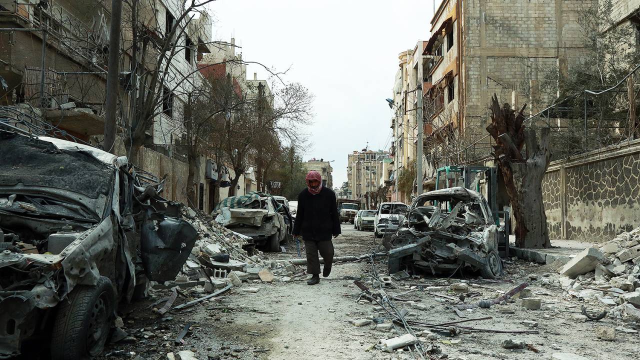 Homem caminha entre veículos destruídos e ruínas de prédios, na cidade de Douma, na Guta Oriental - 25/02/2018
