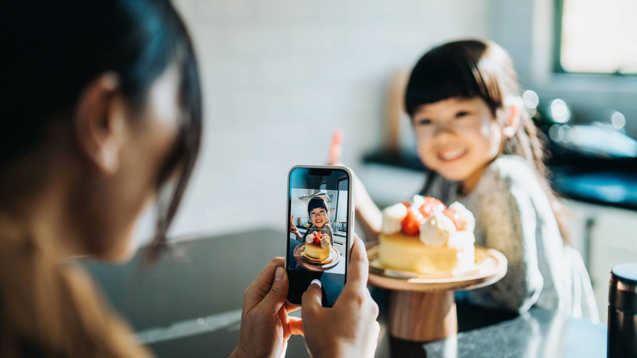 Cropped shot of young Asian mother celebrating 5th birthday with her lovely daughter, taking pictures with smartphone of the birthday girl holding a number 5 candle in front of the birthday cake. Birthday celebration and party theme