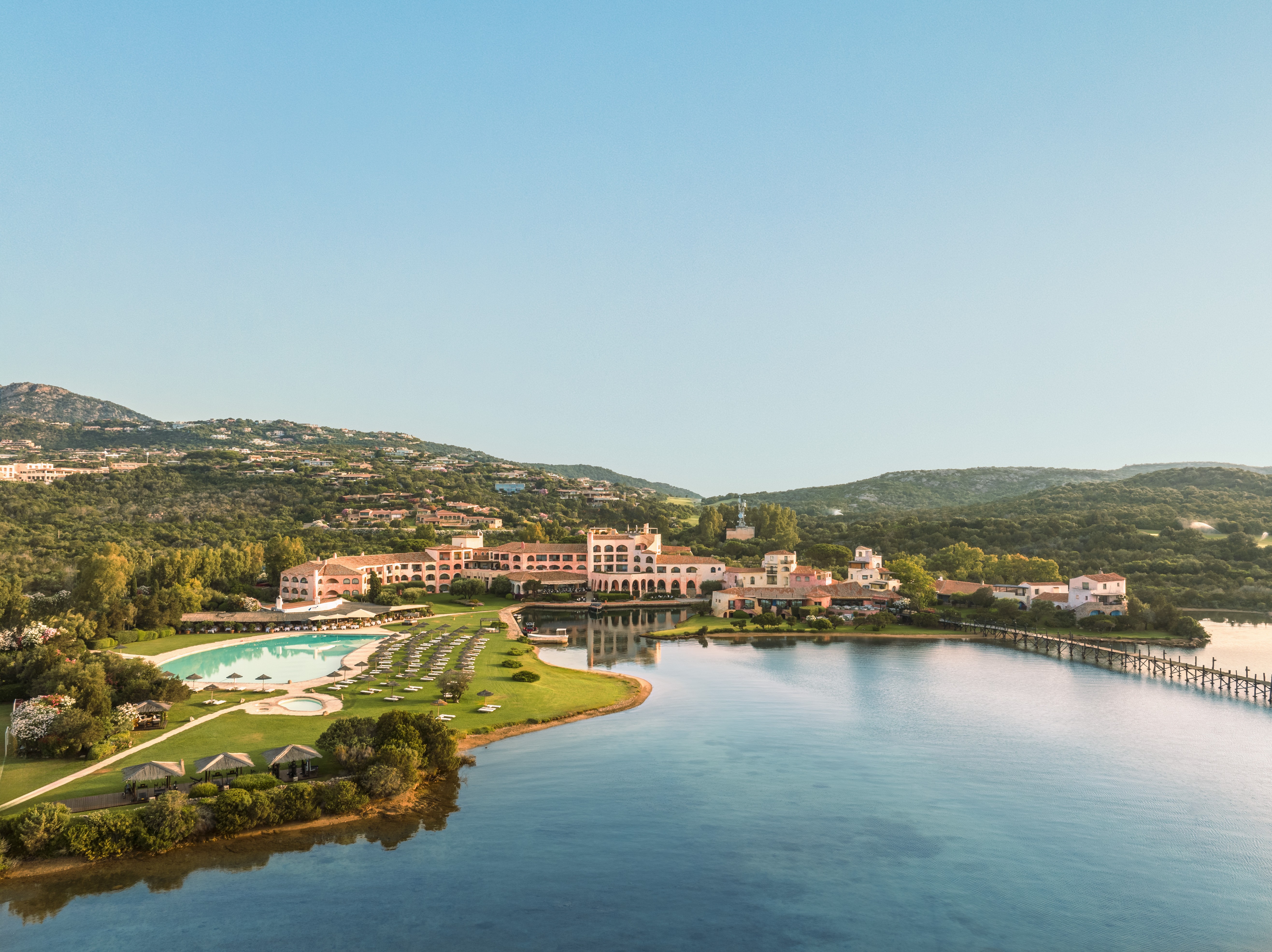 Vista aérea de um resort luxuoso com edifícios cor de pêssego, piscinas azuis e espreguiçadeiras em gramados verdes, à beira de um lago. Ao fundo, montanhas cobertas por vegetação e casas, sob um céu azul claro. Uma ponte de madeira atravessa o lago à direita