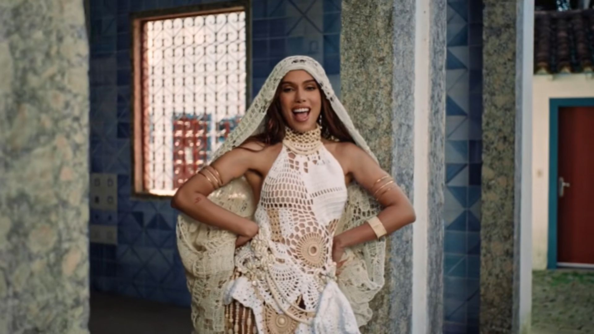 Anitta sorrindo, usando um vestido de crochê branco e bege, com um véu cobrindo a cabeça e ombros, braços na cintura, em um cenário com paredes de azulejos azuis e uma porta vermelha ao fundo