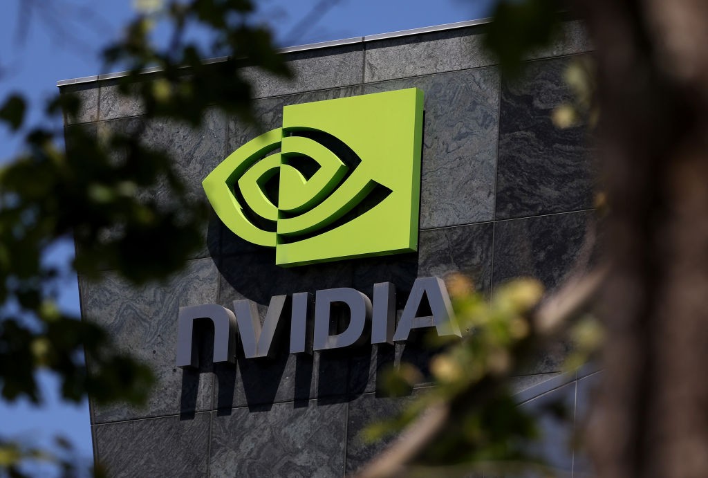 O logotipo da Nvidia, um olho estilizado em verde-limão e preto, está fixado em uma parede de mármore escuro, com o nome da empresa em letras cinzas abaixo. Folhas de árvores em primeiro plano desfocam a visão, e o céu azul é visível acima
