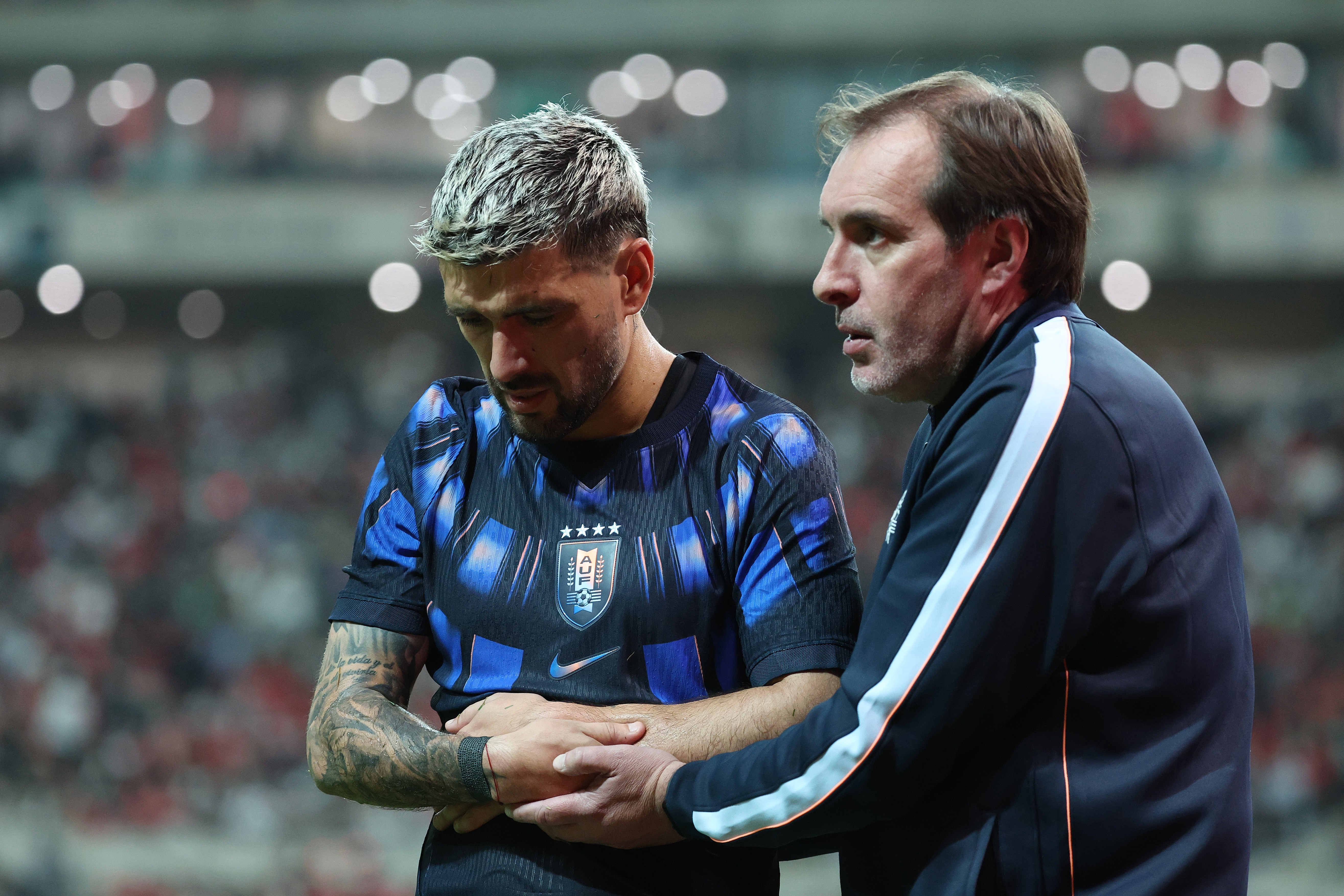 Um jogador de futebol com cabelo grisalho e barba, vestindo camisa azul escura com detalhes azuis brilhantes, segura o abdômen enquanto um homem de meia-idade com jaqueta azul marinho e listras brancas o observa, aparentemente preocupado, em um estádio com luzes desfocadas ao fundo