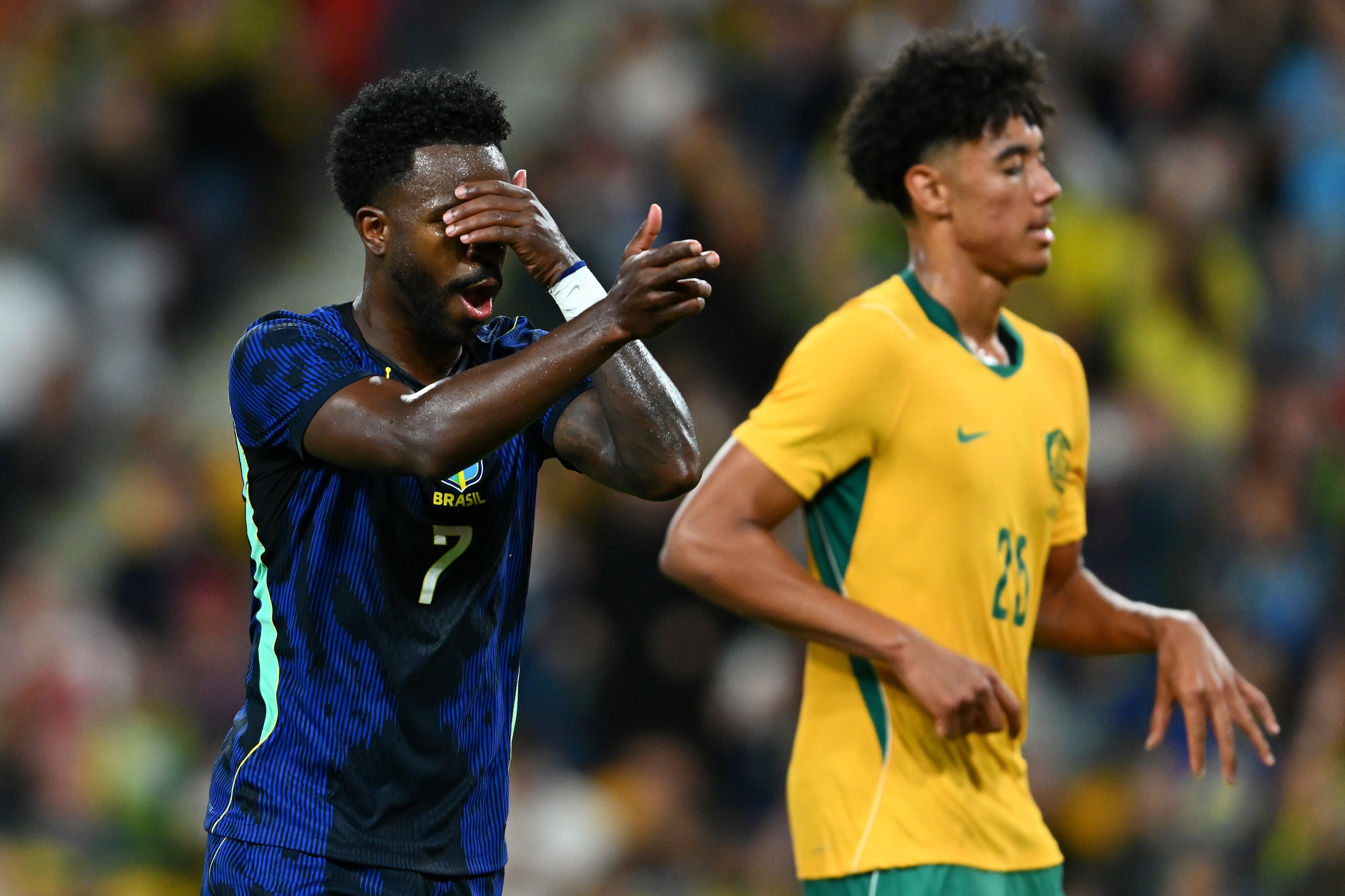 Dois jogadores de futebol em campo, um homem negro de camisa azul com listras pretas e número 7, cobrindo o rosto com a mão direita e gesticulando com a esquerda, e outro homem de camisa amarela e verde, com o número 25, olhando para a direita com expressão séria