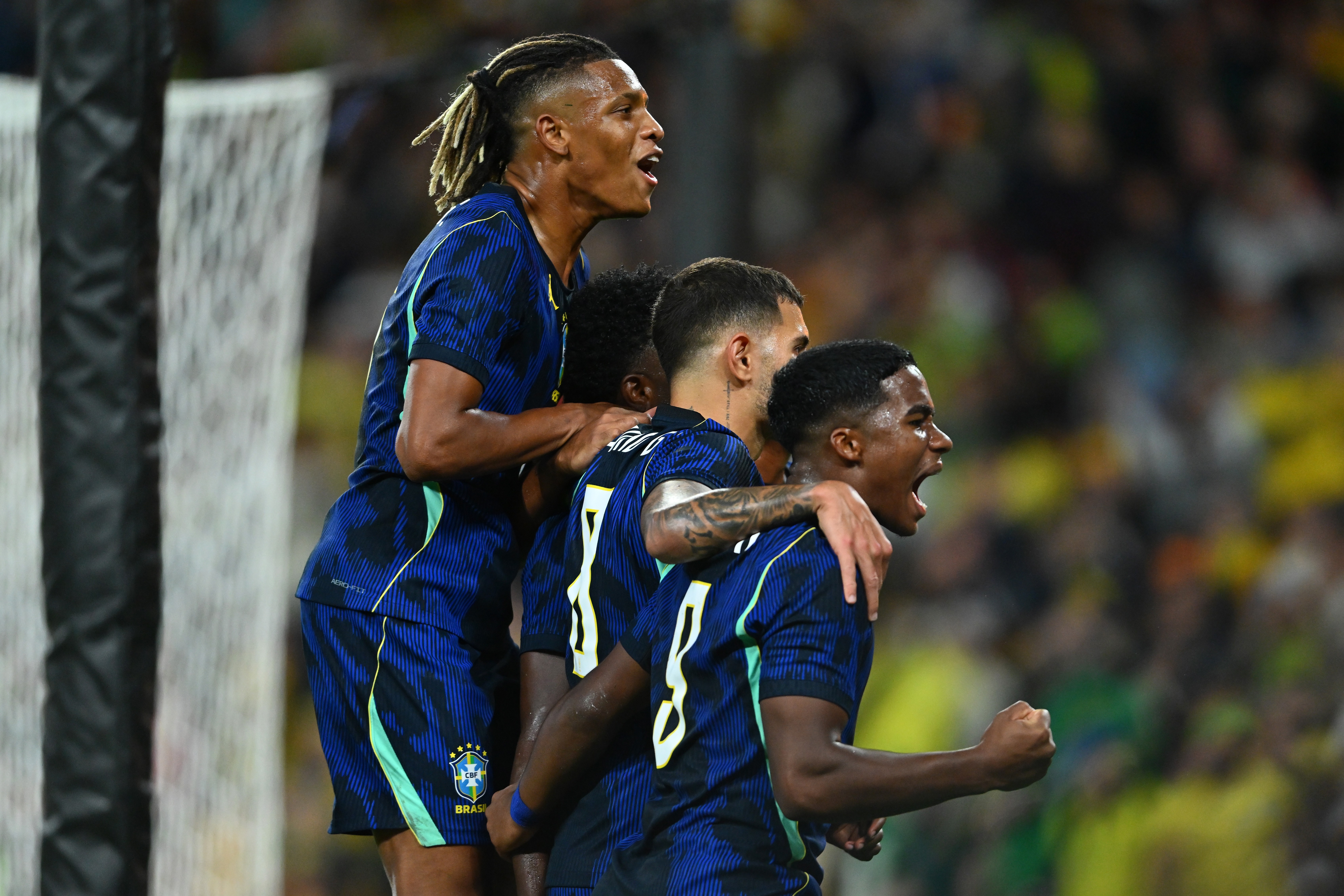 Quatro jogadores de futebol brasileiros, vestindo uniformes azuis com detalhes verdes, celebram um gol. O jogador mais alto, com dreadlocks, está à esquerda, com a boca aberta e braços levantados. Os outros três estão mais próximos, com expressões de euforia e braços ao redor uns dos outros, em um campo de futebol com torcida ao fundo