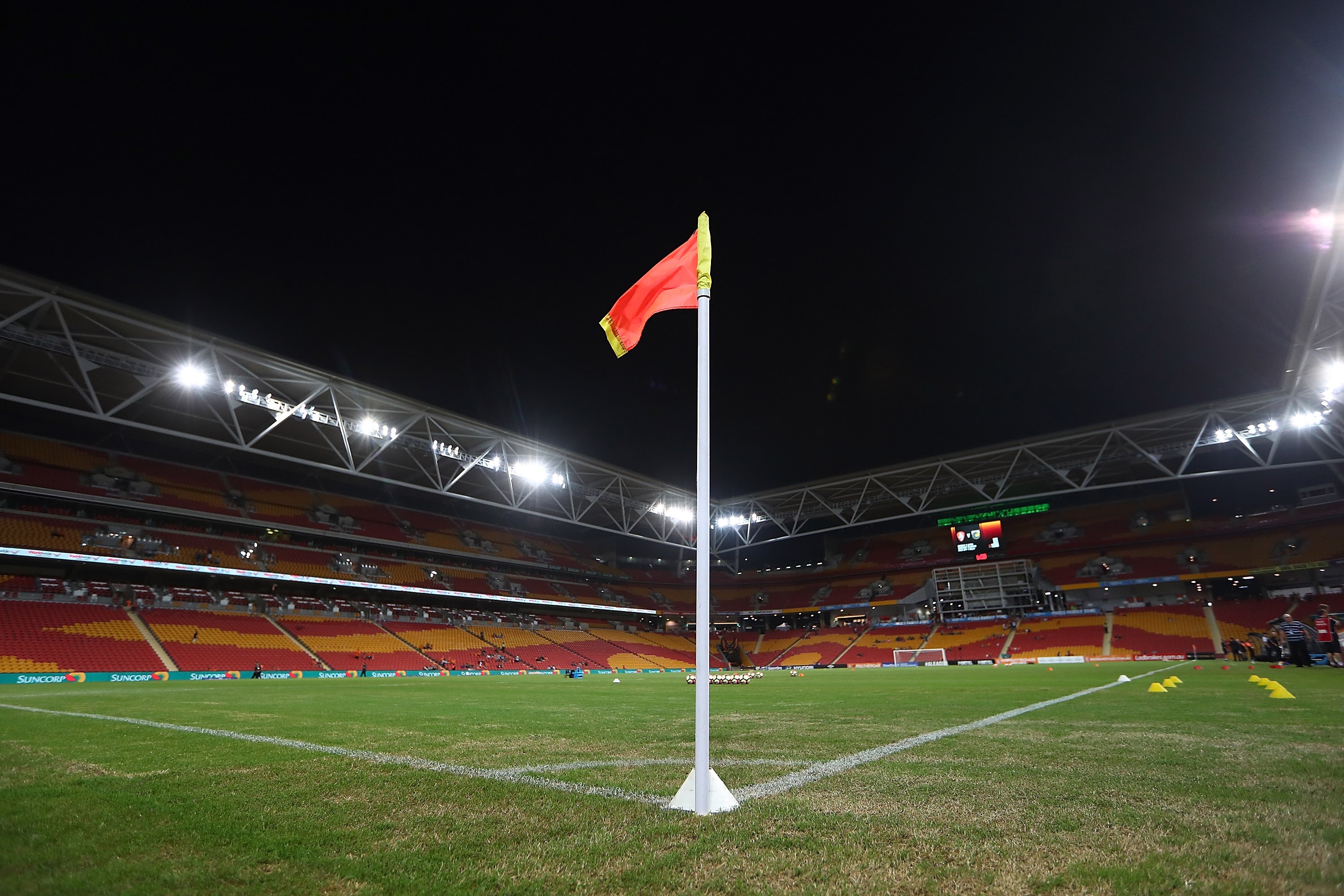 Bandeirinha de escanteio vermelha e amarela em campo de futebol gramado, com arquibancadas vazias em tons de vermelho e amarelo, iluminadas por holofotes sob céu noturno