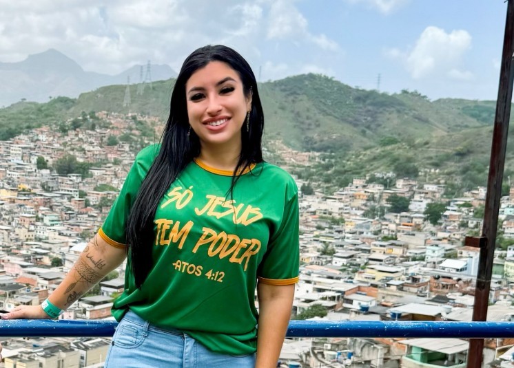 Mulher sorridente de cabelos longos e pretos, vestindo camiseta verde com a frase SÓ JESUS TEM PODER ATOS 4:12 em amarelo, apoiada em um corrimão azul. Ao fundo, uma paisagem urbana com casas simples em uma encosta de morro e montanhas sob céu nublado