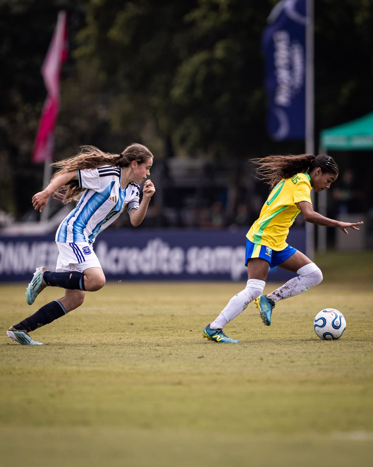 Duas jogadoras de futebol correm em campo gramado. À esquerda, uma atleta de camisa argentina listrada azul e branca persegue a bola. À direita, uma jogadora brasileira de camisa amarela e shorts azuis dribla a bola com o pé direito. Ambas têm cabelos longos e soltos, em movimento. Ao fundo, faixas e árvores desfocadas