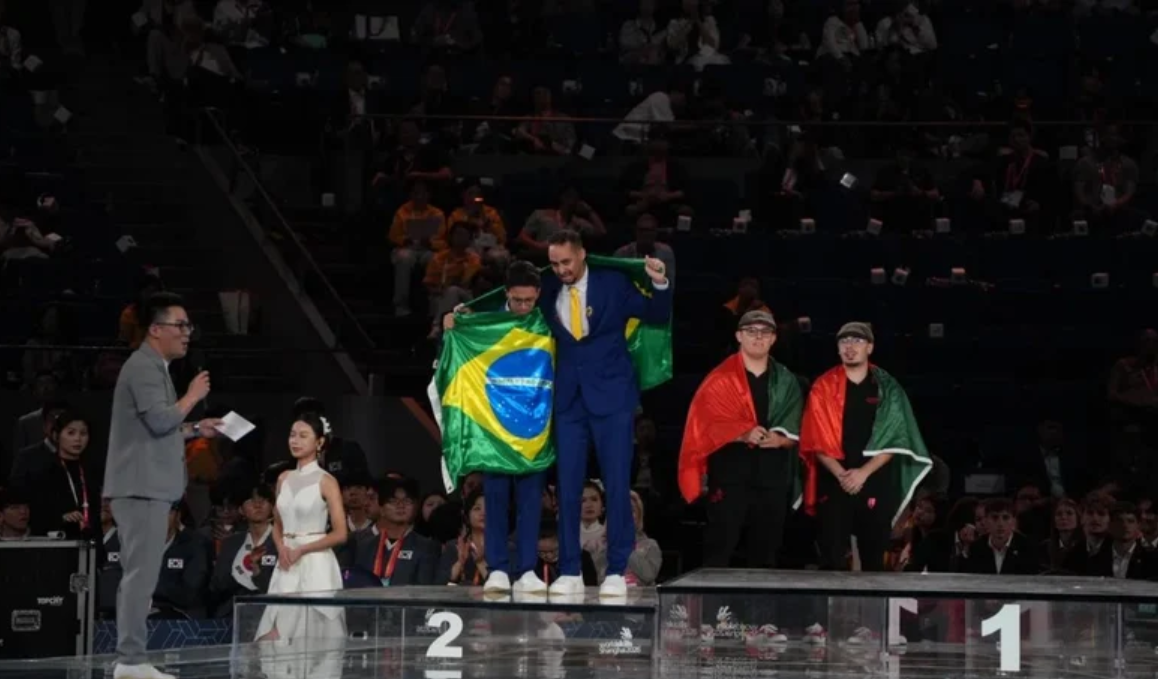 Pódio de premiação com atletas. Dois homens no centro, um de terno azul e outro de camiseta branca, seguram a bandeira do Brasil. À direita, dois homens com bonés e capas vermelhas e verdes. À esquerda, um apresentador de terno cinza fala ao microfone, ao lado de uma mulher de vestido branco. O público lota as arquibancadas escuras ao fundo